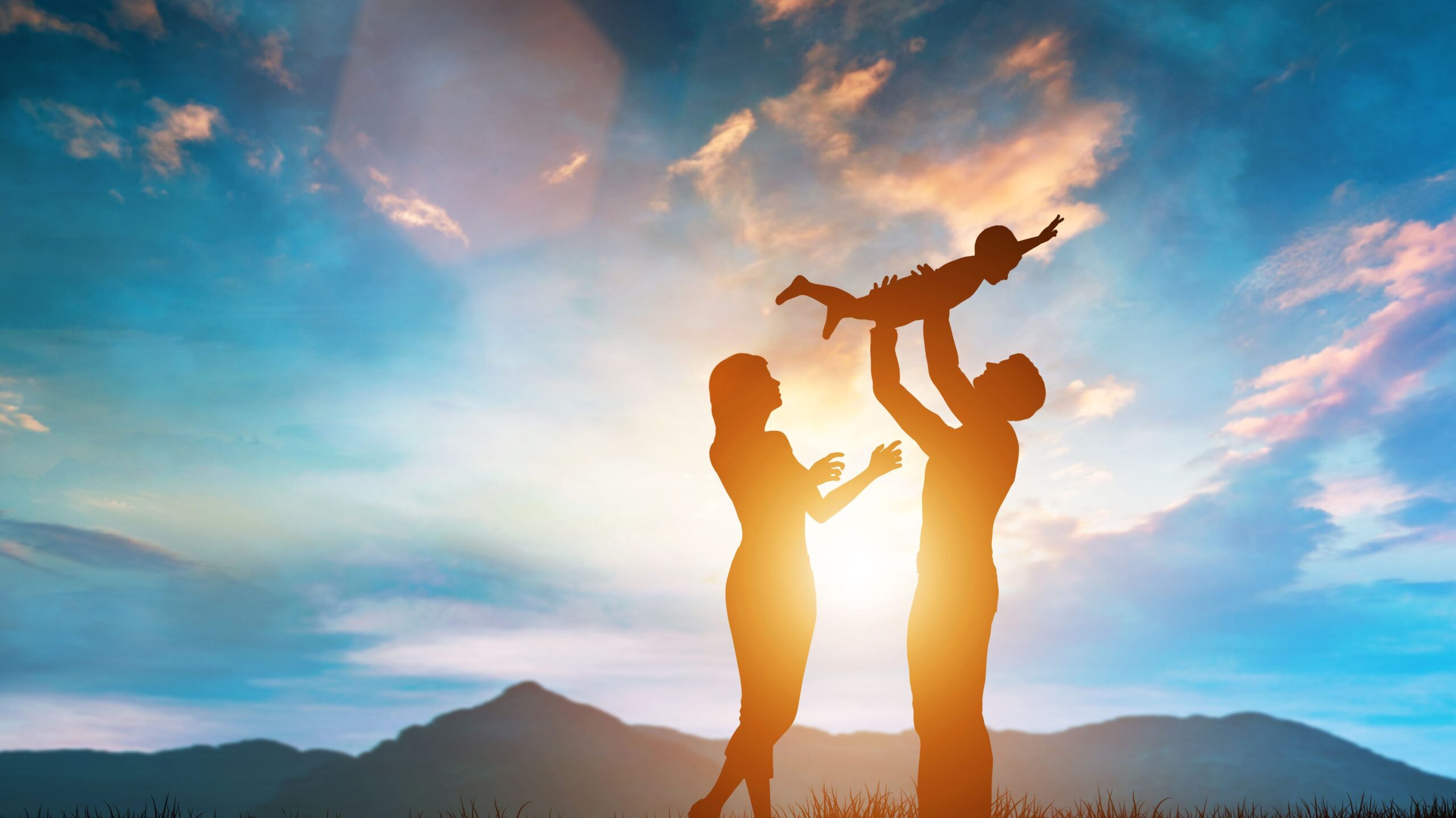 Dad lifting child up in front of sunset next to Mom.