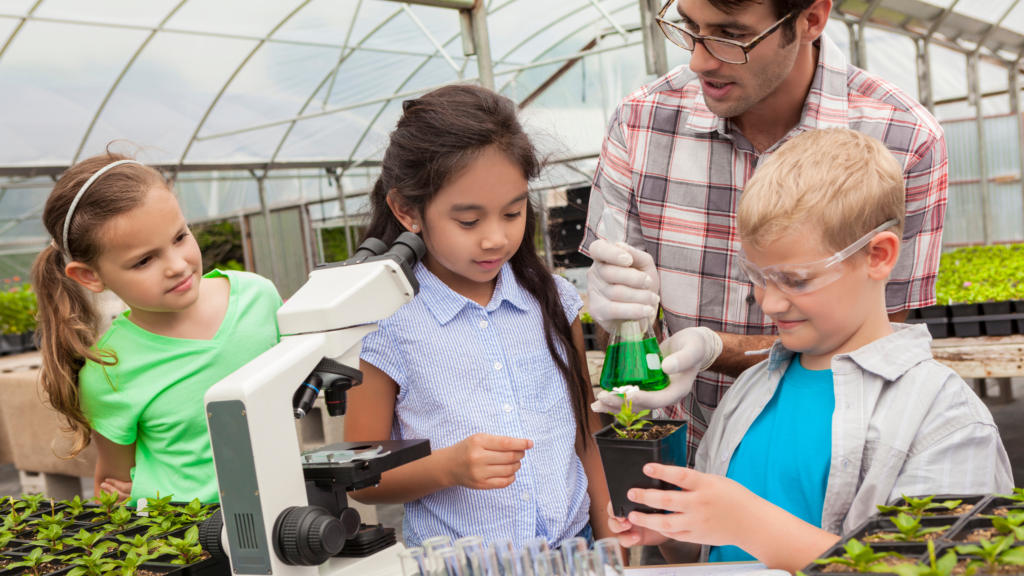 Kids in Greenhouse with scientist looking through microscope holding green vial.